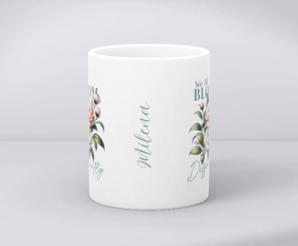 Weiße Keramiktasse mit Blumenmotiv, dem Zitat „We All Bloom Differently“ und personalisiertem Namen „Milena“, auf hellem Hintergrund präsentiert.